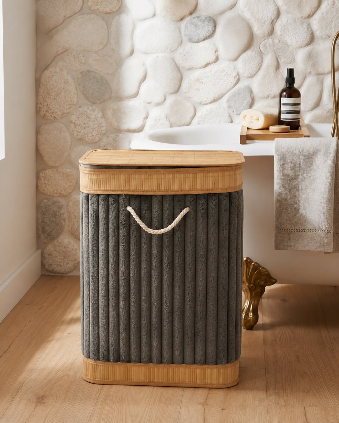 A ribbed gray fabric laundry hamper with a bamboo lid and base, featuring a rope handle, set in a bathroom next to a white tub.