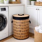 A tall cylindrical woven laundry hamper with a bamboo-style body, black patterned accents, and a black handle, placed in a bright laundry room next to a washing machine.