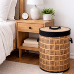 A woven cylindrical laundry hamper with a black handle and trim, placed next to a wooden nightstand in a bedroom setting.