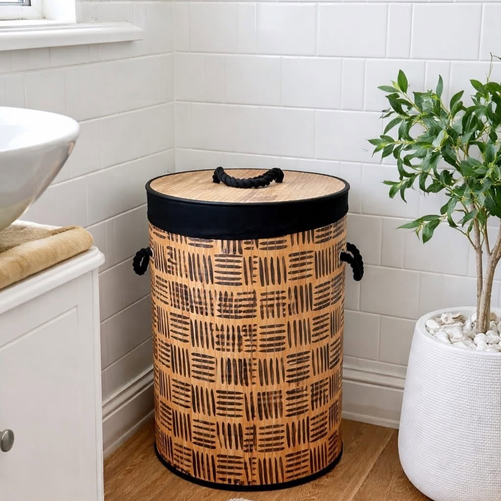 A cylindrical woven trash can with a bamboo-style top and black trim, placed in a bright bathroom corner next to a white sink and a potted plant.