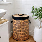 A cylindrical woven trash can with a bamboo-style top and black trim, placed in a bright bathroom corner next to a white sink and a potted plant.