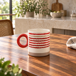 A ceramic mug with a red handle and red wavy horizontal stripes on a white body, sitting on a wooden table in a bright kitchen setting.